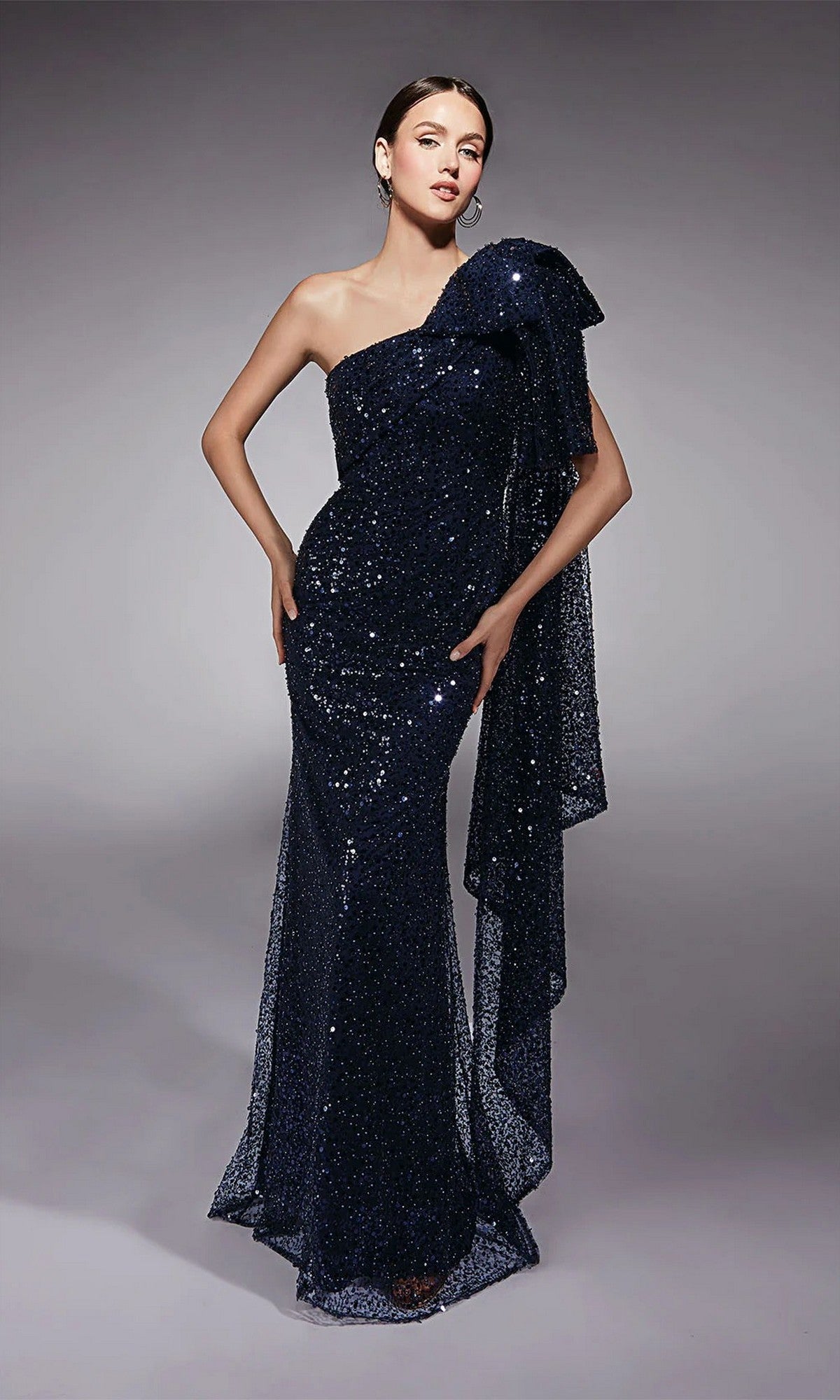 A woman models the Alyce 27771 Long Formal Dress in navy—a floor-length, one-shoulder sequin gown with draped side detail—standing against a plain gray background, highlighting its elegant evening style.